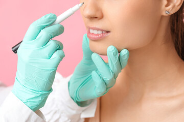 Doctor in protective gloves drawing marks on woman's nose for plastic surgery against pink...