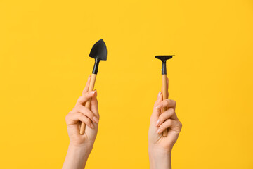 Female hands with gardening shovel and rake on yellow background, closeup
