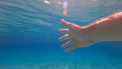 POV UNDERWATER, LENS FLARE: Stunning crystal clear blue Adriatic Sea with a male hand gently moving...