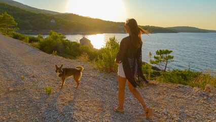 LENS FLARE: Walking of a young tourist with a dog along picturesque gravel road. Cheerful woman is...