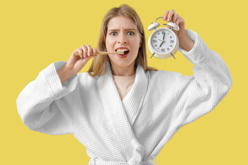 Shocked young woman in bathrobe holding alarm clock and brushing teeth on yellow background