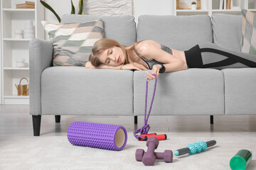 Tired young woman lying on sofa with sports equipment at home