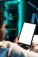 BIPOC man sitting on sofa, reading eBook from tablet in apartment using mockup tablet. Relaxed person enjoying interesting lecture on isolated screen device in neon lit living room