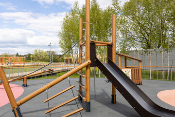 An outdoor playground featuring a wooden climbing frame with a slide, rope ladder, and swing set,...