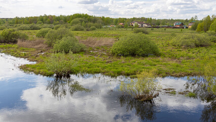  A tranquil wetland scene with reflections of lush greenery and trees in the calm water, bordered by a rural community with houses and overgrown vegetation.