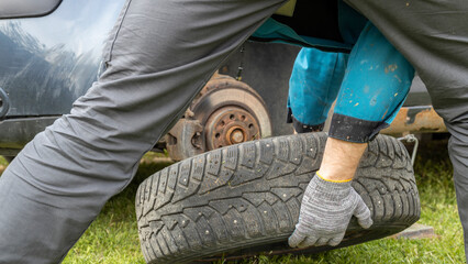 A man changes a tire, holding it with his hands; green grass indicates the timely replacement of...