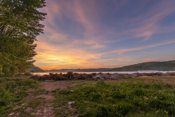 A tranquil sunset over a lake with a mix of orange, blue, and purple skies. The foreground shows a path leading to a pile of wooden logs and debris, with a distant ship on the horizon.