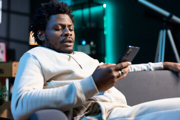 African american man reading messages on smartphone, receiving bad news from friends cancelling...