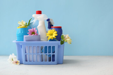 Basket with set of cleaning supplies and spring flowers on white table against blue background