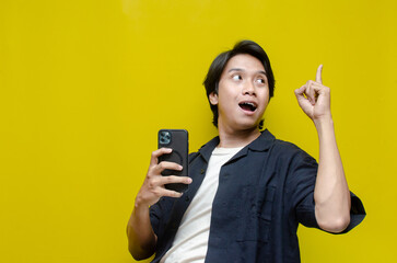 excited young asian man holding phone while dancing and pointing to copy space with full of...
