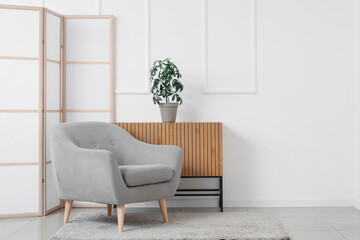 Interior of light living room with grey armchair, chest of drawers and folding screen near white wall