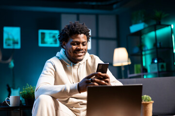 Smiling man at home taking break from work to write messages on phone. Joyous person holding...