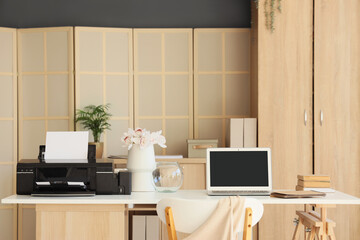 Modern printer with flowers and laptop on desk in office