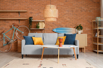 Surfboard, bicycle, cozy sofa and shelving units near brown brick wall in living room