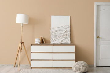 Lamp, picture and chest of drawers near beige wall in stylish room