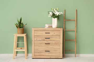 Vase with lily flowers on wooden chest of drawers near green wall in room