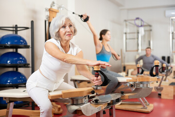 Elderly woman in sportswear stretches her arms and shoulders using pilates machine..