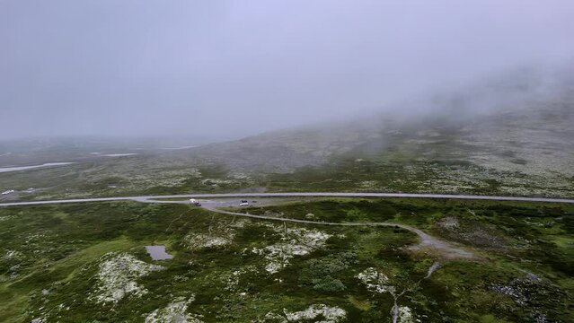 Flying over the green tundra and above a parking lot in Rondane National Park, near Venabygd, Norway, showcasing the misty nature landscape and disappearing into the cloudy mountains
