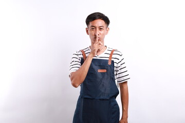 Asian young man wearing apron over white background asking to be quiet with finger on lips. silence...