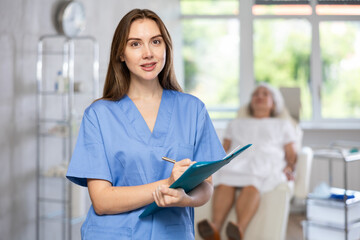 Positive confident female doctor in medical uniform holding clipboard while elderly patient lying on exam table.