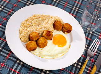 Hearty breakfast setup showing creamy oatmeal with sunny side up egg and savory meatballs on white...