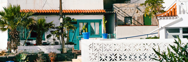 A cozy white house in a cortyhard with a green tropical garden, cacti and palm trees.