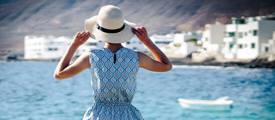 A girl in a blue dress with a hat on her head looks at the beautiful ocean on a sunny day.