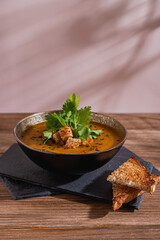close up of golden pumpkin soup with croutons, big leaves of cilantro and sesame seeds on top over slate boards and wooden rustic table with toasted bread triangles