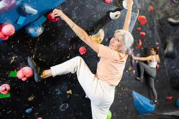 Joyful concentrated senior woman busy with her hobby bouldering, climbing on artificial wall in...