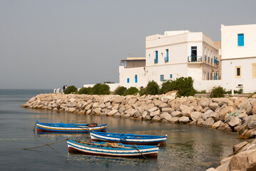 Traditional blue fishing boats floating on calm waters of Mediterranean Sea by stony shoreline, in front of Mahdia picturesque white buildings with vibrant blue windows, under clear sky