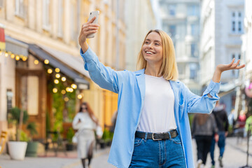 Cheerful woman blogger taking selfie on smartphone, communicating video call online with subscribers or family friends, recording stories for social media vlog, waving hello. Lady on urban city street