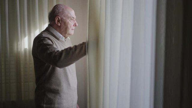 A senior man gazes out a window, sunlight streaming through sheer curtains and illuminating the room, reflecting a moment of contemplation.