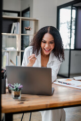 A woman is sitting at a desk with a laptop and smiling. She is happy and excited about something on the screen