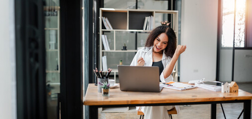 A woman is sitting at a desk with a laptop and a stack of papers. She is smiling and she is happy