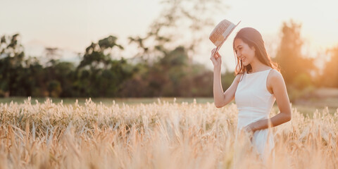 A woman is standing in a field of tall grass, wearing a straw hat and a white dress. She is smiling...