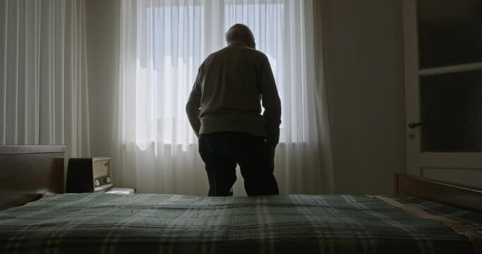 An elderly man sits alone on a bed in a sunlit room, staring out a window with sheer curtains.