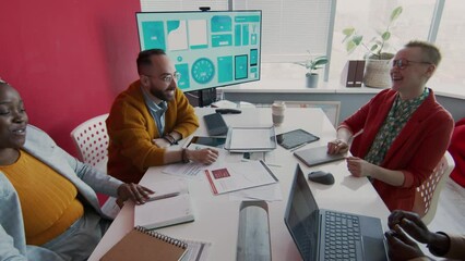 Medium shot of creative multinational project team sitting together around table in meeting room, discussing new marketing campaign then joining hands together for motivation and laughing