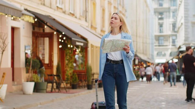 Young blonde woman tourist with luggage suitcase exploring checking paper map search a way direction while traveling abroad. Lady girl walking on urban summer city street. Town lifestyles outdoors