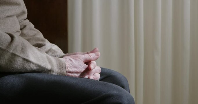 A close-up of a senior mans hands clasped together, conveying anxiety, while sitting in a room with light-colored curtains.