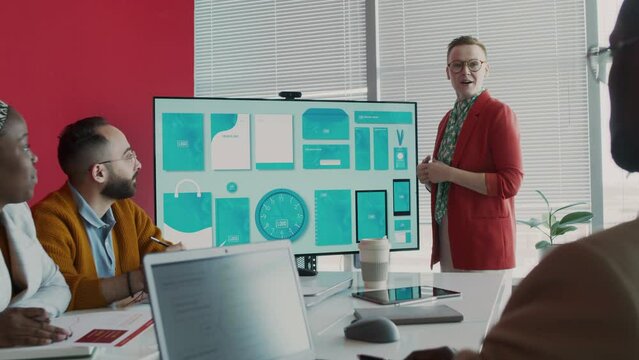 Medium shot of inspirational Caucasian female product manager presenting new merchandise design for advertising campaign on multimedia screen at meeting in front of diverse colleagues