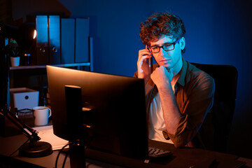Young businessman calling on smartphone to customer or coworker, looking at computer screen of...