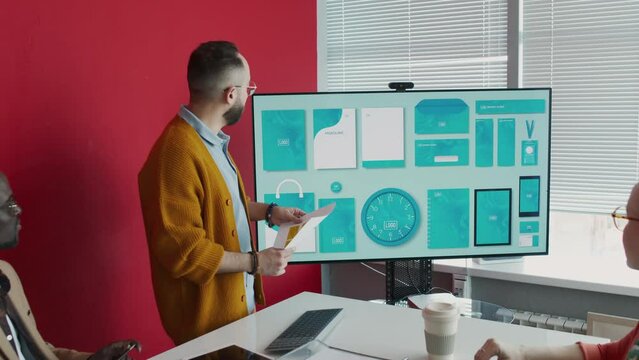 Medium zoom-out shot of biracial brand manager presenting draft layout of new stationery for promotion campaign in front of creative team, on multimedia screen, multiethnic colleagues watching