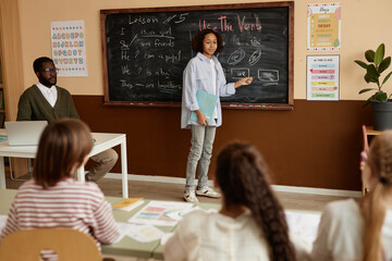 Modern Black teenager attending English language class at school standing at blackboard doing...