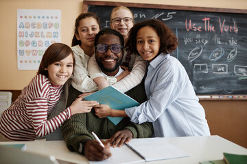 Group portrait of ethnically diverse students standing in classroom hugging their teacher and...