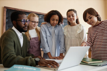 Young African American teacher and group of multi-ethnic boys and girls looking at laptop screen in...