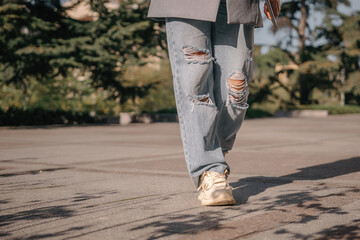 A woman is walking down a sidewalk wearing ripped jeans and a gray jacket. The image conveys a...