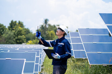 A man in a blue shirt and white hat is standing in a field of solar panels. He is holding a...