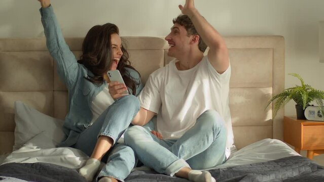 Caucasian couple sitting on bed bedroom looking at screen smartphone mobile phone man woman enjoyment cellphone homeowner good news victory success achievement emotions celebrating surprise excitement