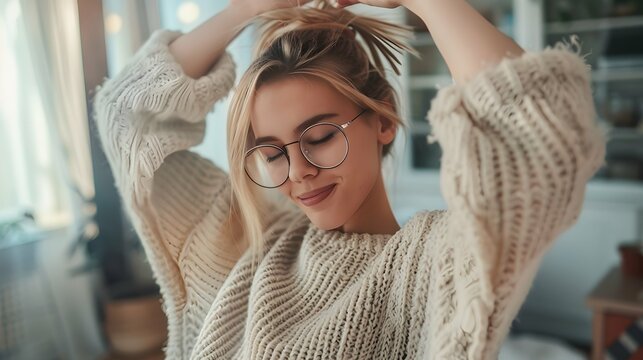 Funny young fair-skinned woman dancing with her eyes closed alone at home in daytime. Blonde with hair pulled back wears translucent glasses and oversized sweater. Freedom and happy lifestyle concept