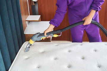 A person in a purple jumpsuit is cleaning a mattress with a steam cleaner